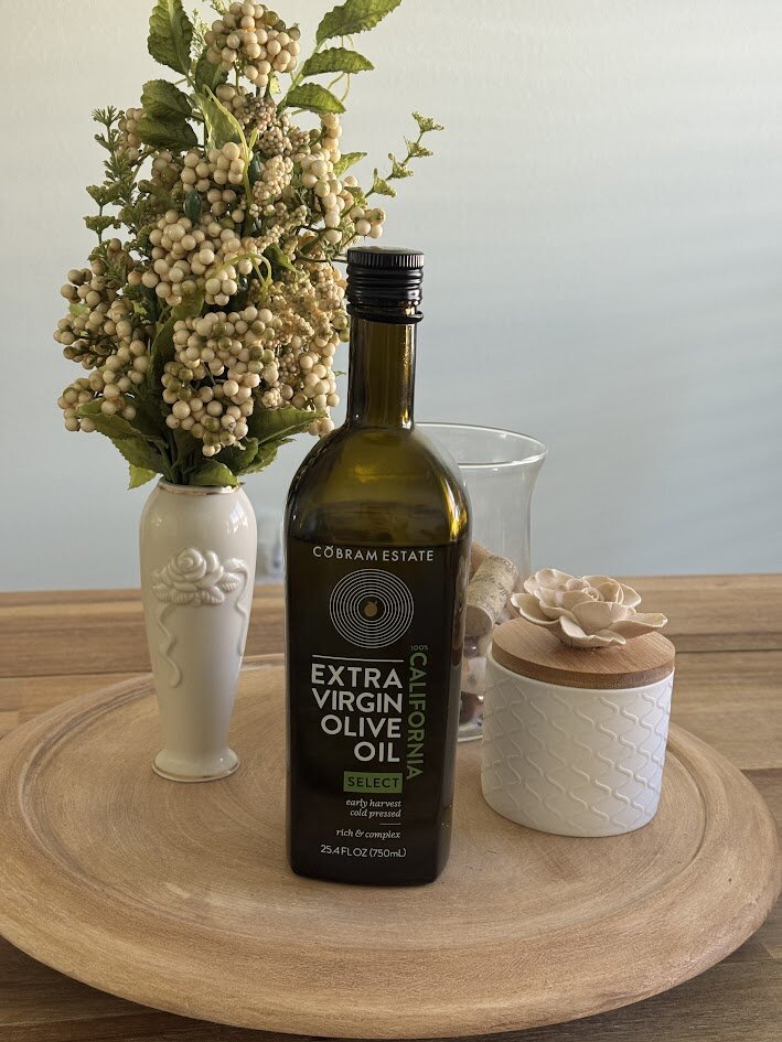 A bottle of Cobram Estate Extra Virgin Olive Oil, Select, on a wooden tray beside a cream vase of berries and a white lidded jar